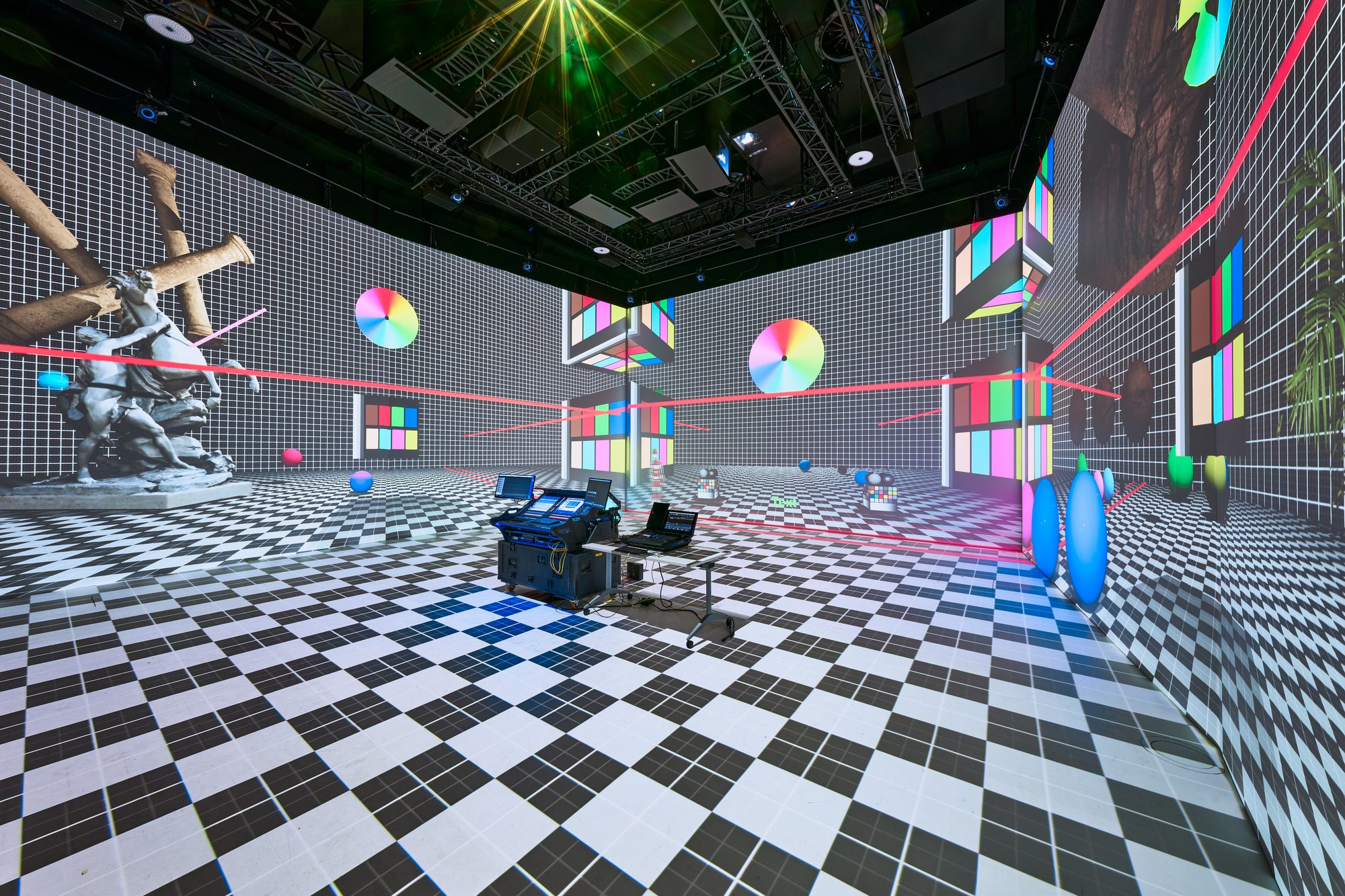 Inside the hexagonal X-Reality-Lab: a checkerboard floor and colour test charts projected across the walls, with a windmill, a sculpture and an equipment cart in the room.