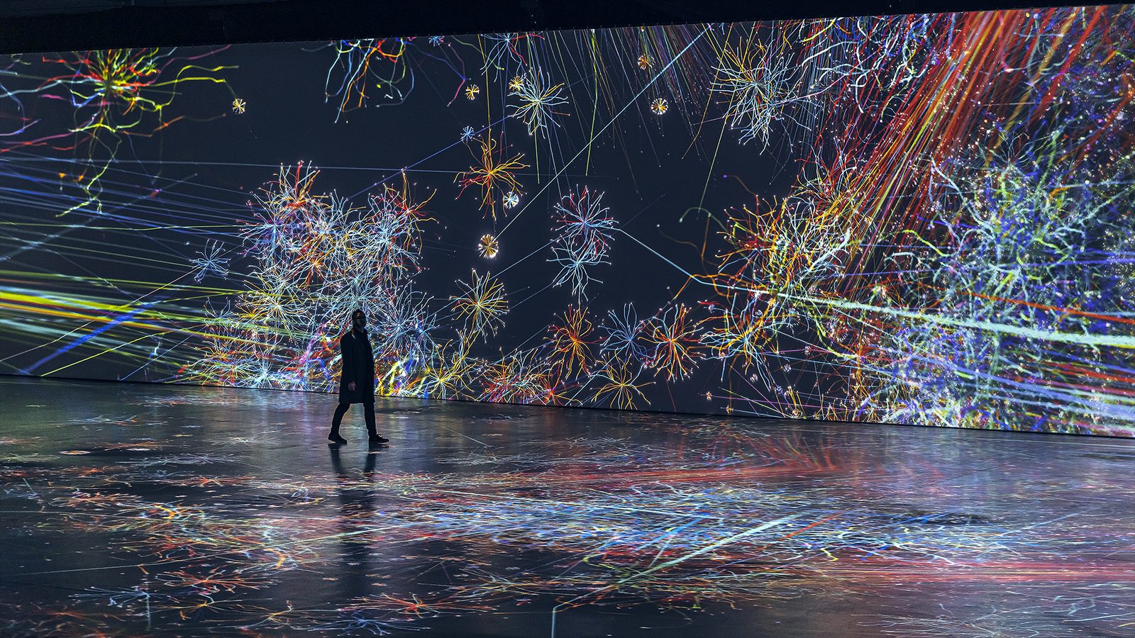 Visitors silhouetted in a vast hall filled with drifting coloured light particles from Nohlab’s Journey.